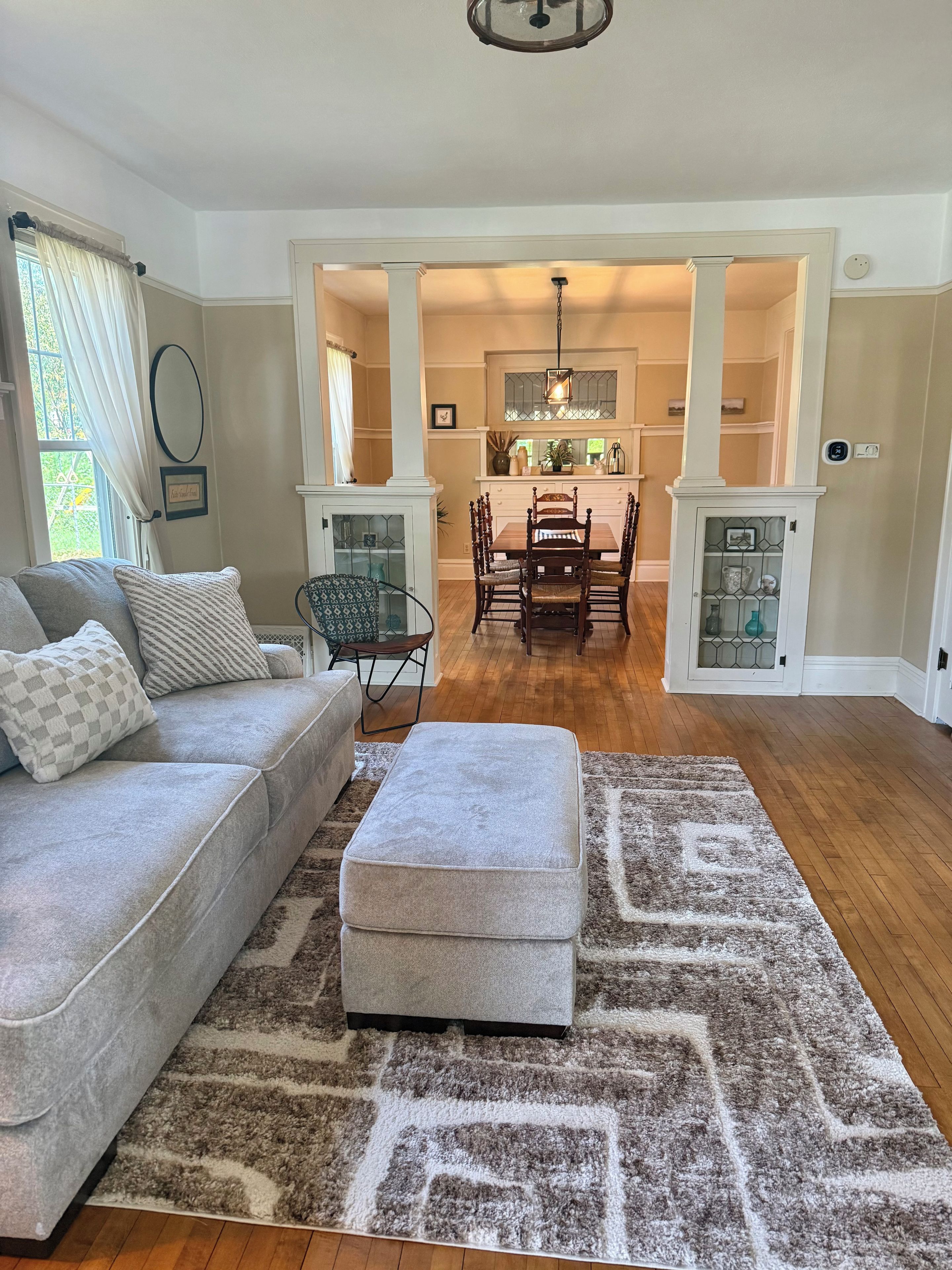 A living room with a grey sofa, ottoman, and patterned rug. A dining area with a wooden table and chairs is visible in the background.
