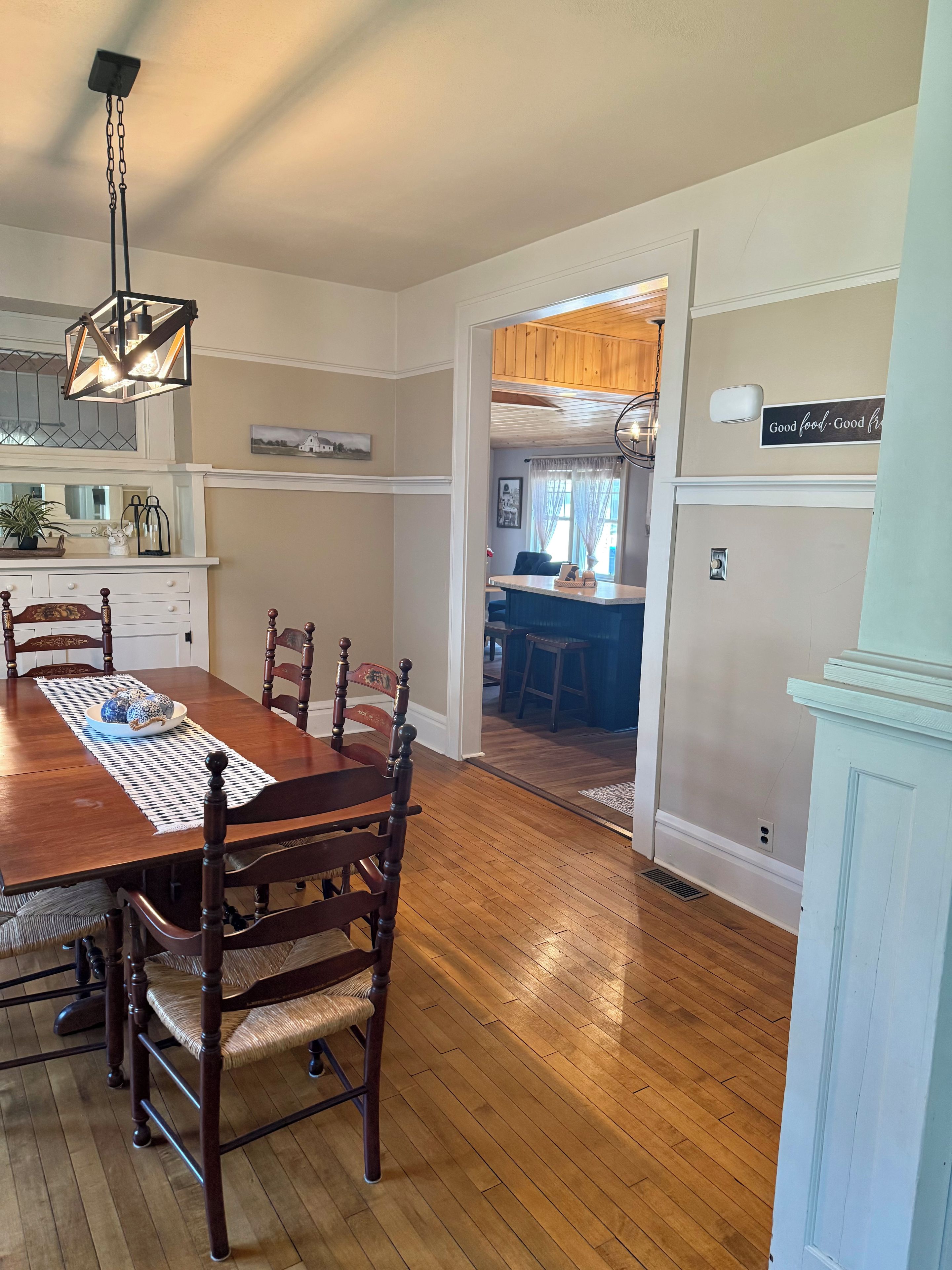 A dining room with a wooden table and chairs, a kitchen in the background, and a sign on the wall.
