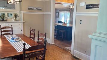 A dining room with a wooden table and chairs, a kitchen in the background, and a sign on the wall.