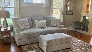 A living room with a grey sectional sofa, an ottoman, a patterned rug, a side table with a lamp, and a chair.