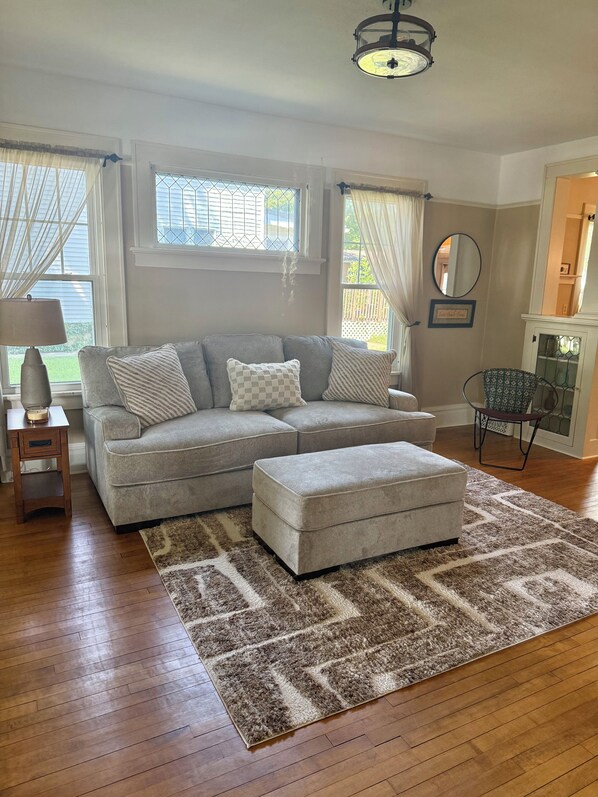 A living room with a grey sectional sofa, an ottoman, a patterned rug, a side table with a lamp, and a chair.