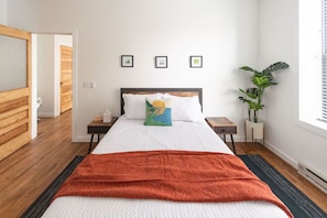 A bedroom with a bed, bedside tables, a window, a plant, and framed pictures on the wall.