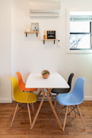 Un pequeño comedor con una mesa blanca y cuatro sillas de colores. Hay un cartel en la pared sobre la mesa y un equipo de aire acondicionado montado en la pared.
