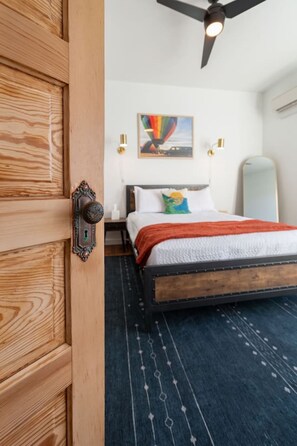 A bedroom with a wooden door, a bed with white bedding and a colorful pillow, a ceiling fan, and a wall-mounted light fixture.