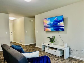 Ein Wohnzimmer mit einem an der Wand befestigten Flachbildfernseher, einer weißen Unterhaltungsanlage und einem braunen Ledersofa mit blauen Kissen.