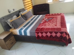 A neatly made bed with a patterned bedspread and pillows, a bedside table with drawers, and a window with blinds. - A Perfect Home for the Family 3 Bed , (Accra Hills)