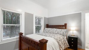Un dormitorio con una cama de madera, dos ventanas con persianas, una lámpara de noche y una mesita de noche.