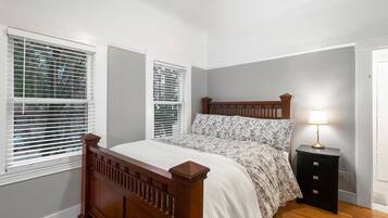 A bedroom with a wooden bed, two windows with blinds, a bedside table with a lamp, and a door.