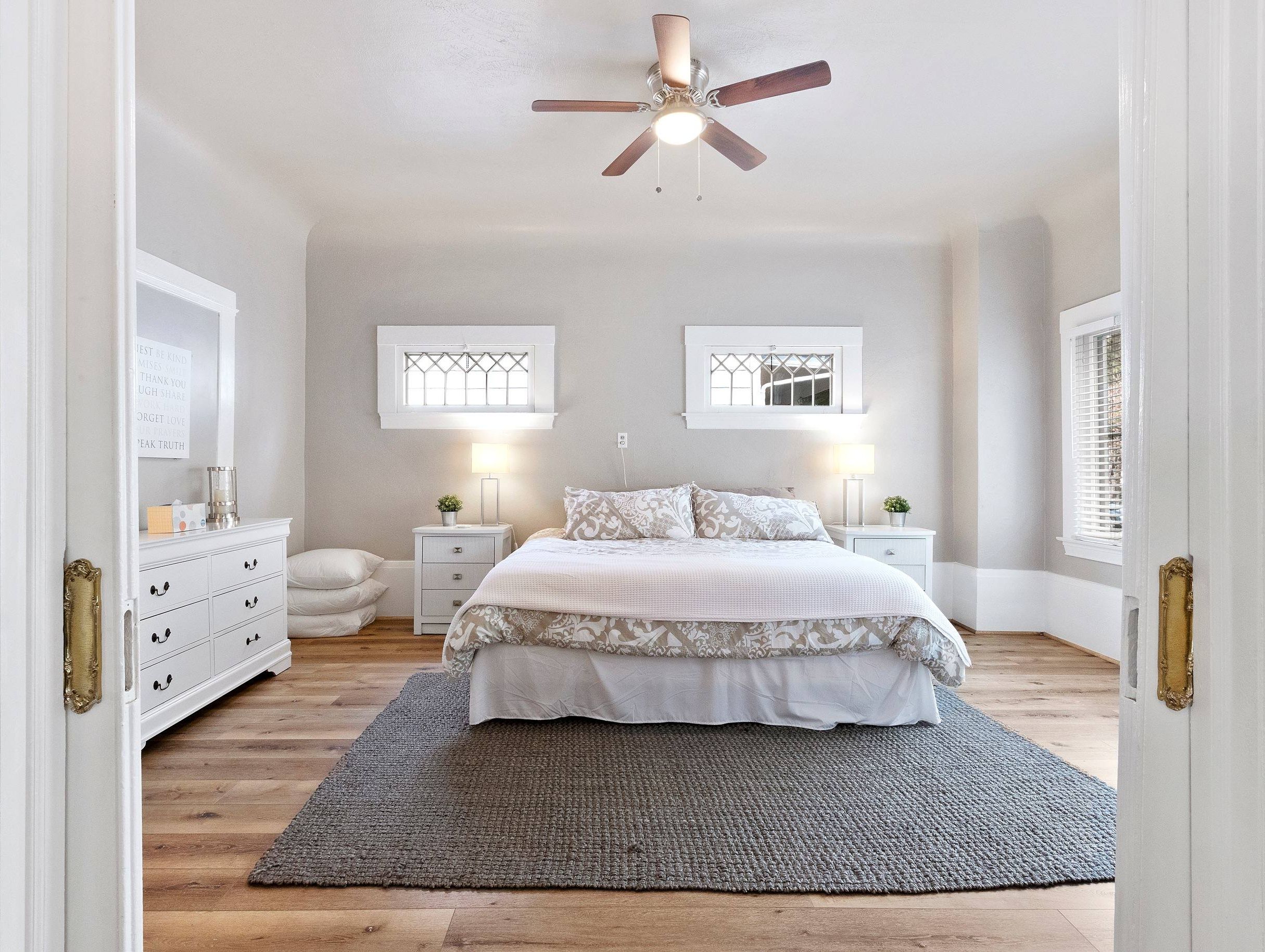 A bedroom with a bed, bedside tables, a ceiling fan, and two windows with shutters.