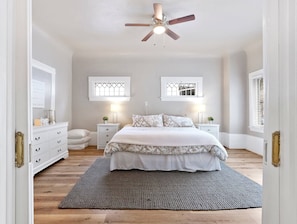A bedroom with a bed, bedside tables, a ceiling fan, and two windows with shutters.