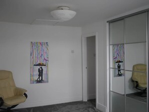 Een kamer met een beige stoel, een glazen scheidingswand en twee kunstwerken aan de muur waarop een stel staat afgebeeld onder een kleurrijke paraplu.