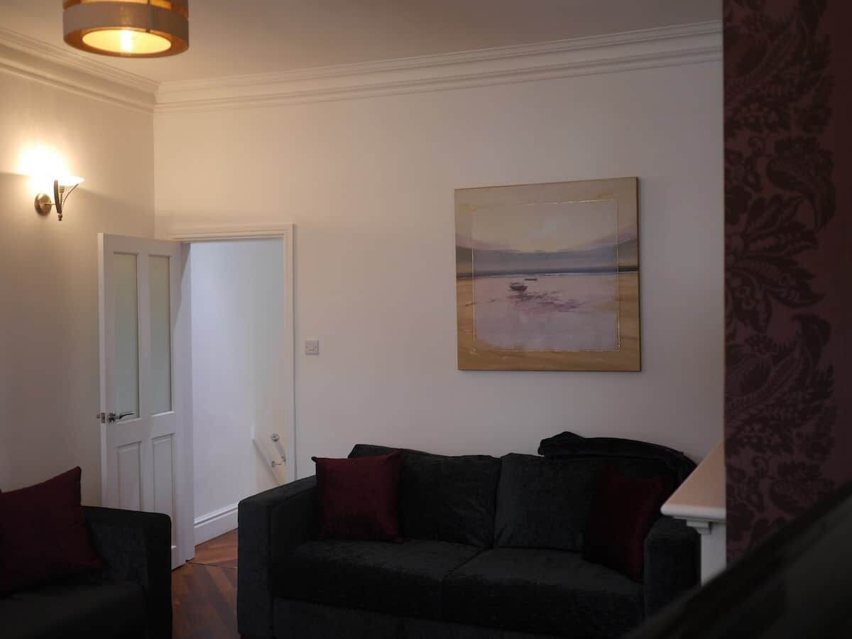 Ein Wohnzimmer mit einem dunkelgrauen Sofa, einem Gemälde an der Wand und einer Tür zu einem anderen Raum.