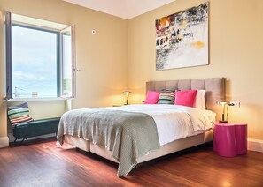 A bedroom with a large bed, a window, a painting, and a bedside table.