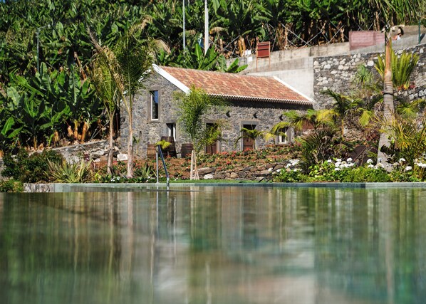 A stone house with a tiled roof, surrounded by lush vegetation and a calm water body.