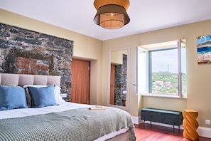 Un dormitorio con pared de piedra, una cama con almohadas, un banco, una ventana con vistas y una lámpara colgante.