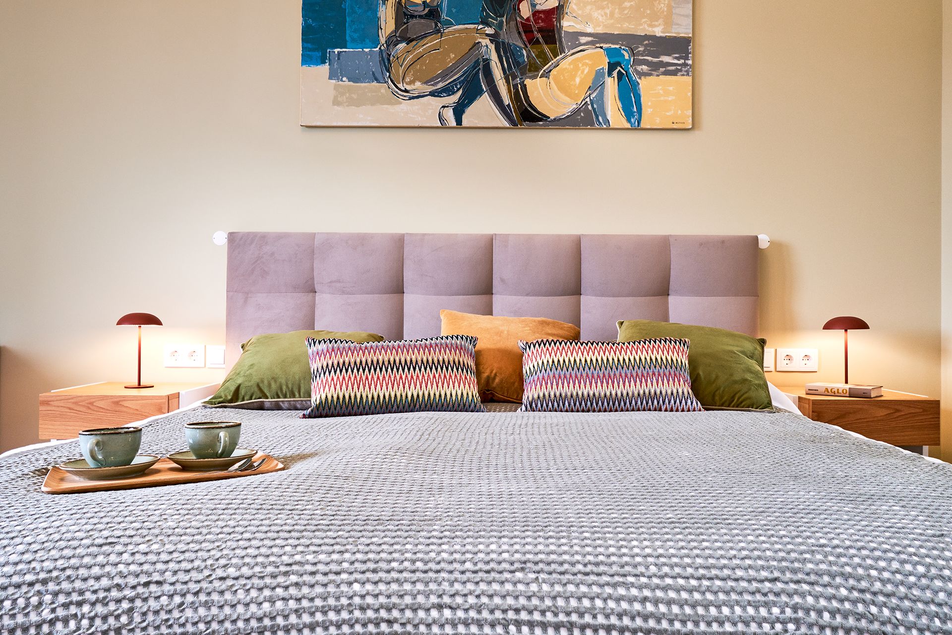 Una cama bien tendida con una colcha estampada, dos almohadas, una bandeja con tazas y un cuadro abstracto colgado en la pared.
