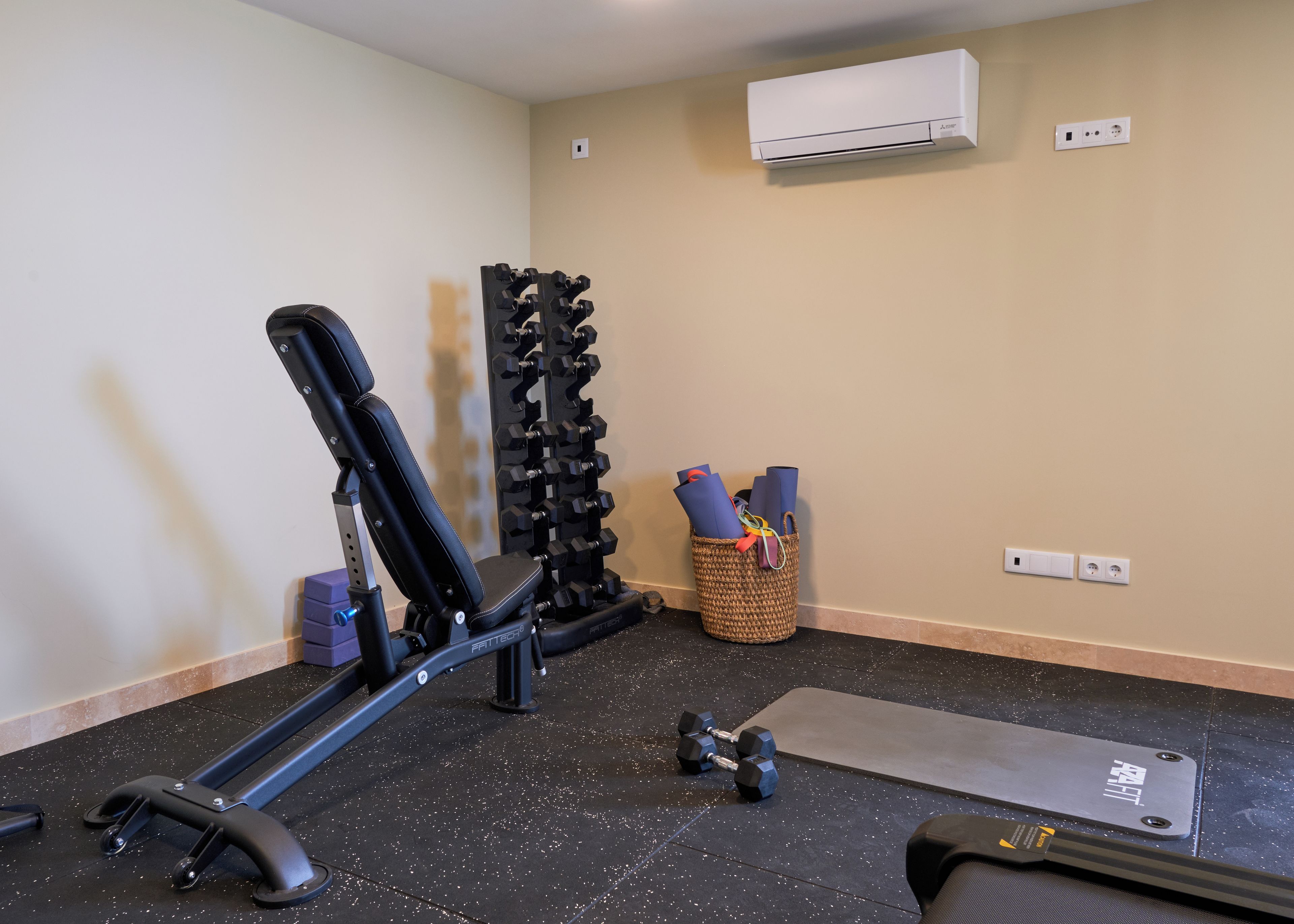 Ein Fitnessraum mit einer Hantelbank, Kurzhanteln, einer Yogamatte und einer Wandklimaanlage.