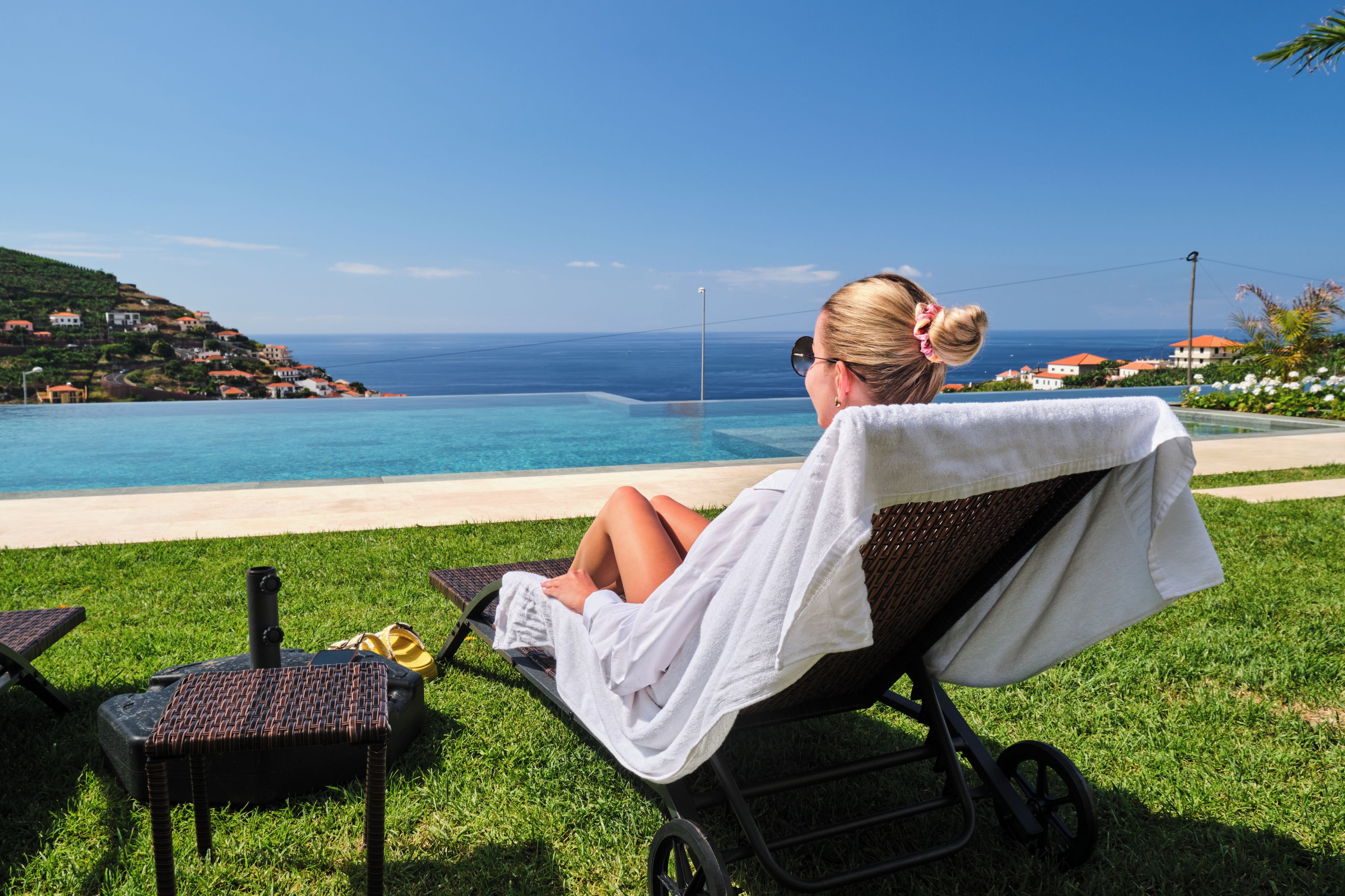 Une personne se détend sur une chaise longue, une serviette à portée de main, et contemple un océan bleu limpide ainsi qu’un paysage vallonné parsemé de bâtiments.