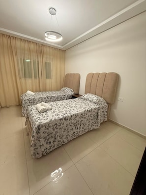 A hotel room with two beds, a ceiling light, a window with curtains, and a tiled floor.