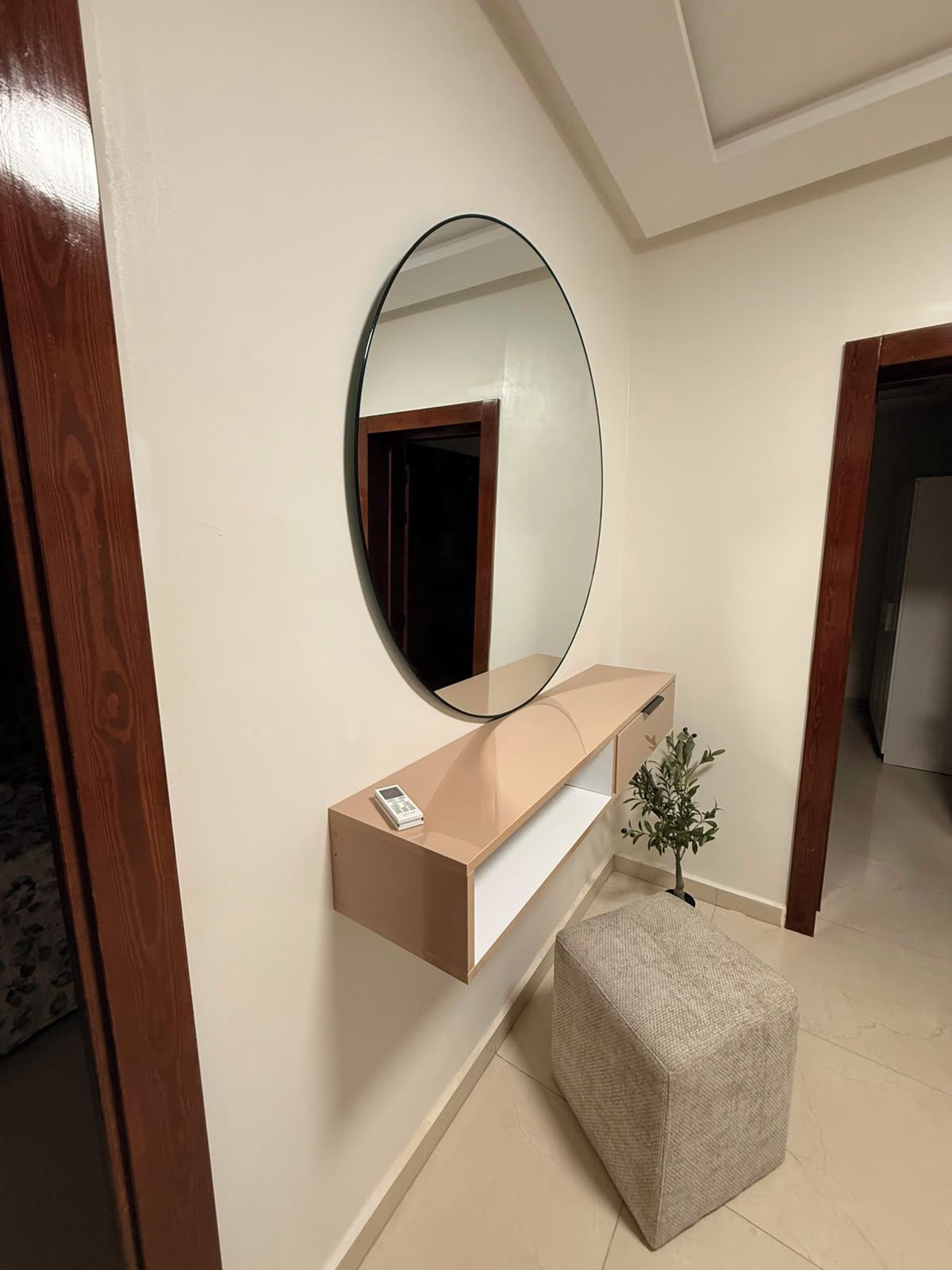 Una habitación con un espejo de marco de madera, una consola de mesa beige, una pequeña planta en maceta y un otomano texturizado.