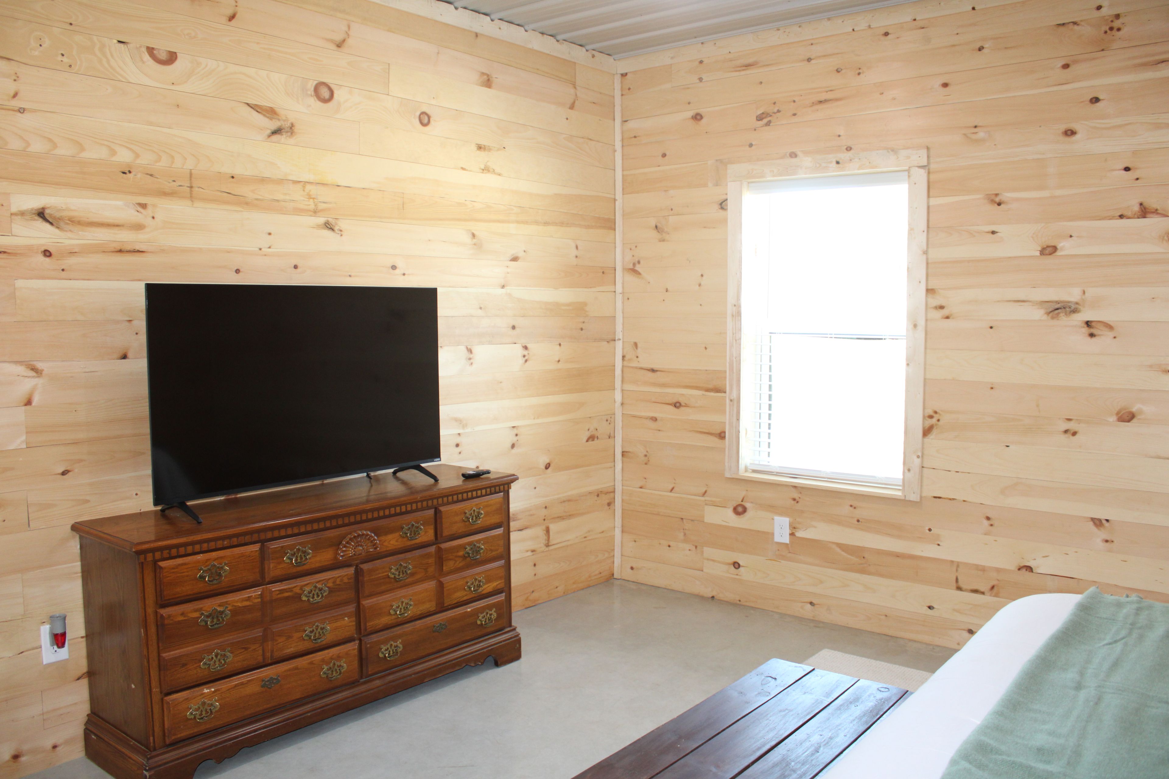 Quarto com paredes de madeira, cômoda de madeira com gavetas, televisão de tela plana em cima e janela com persianas.
