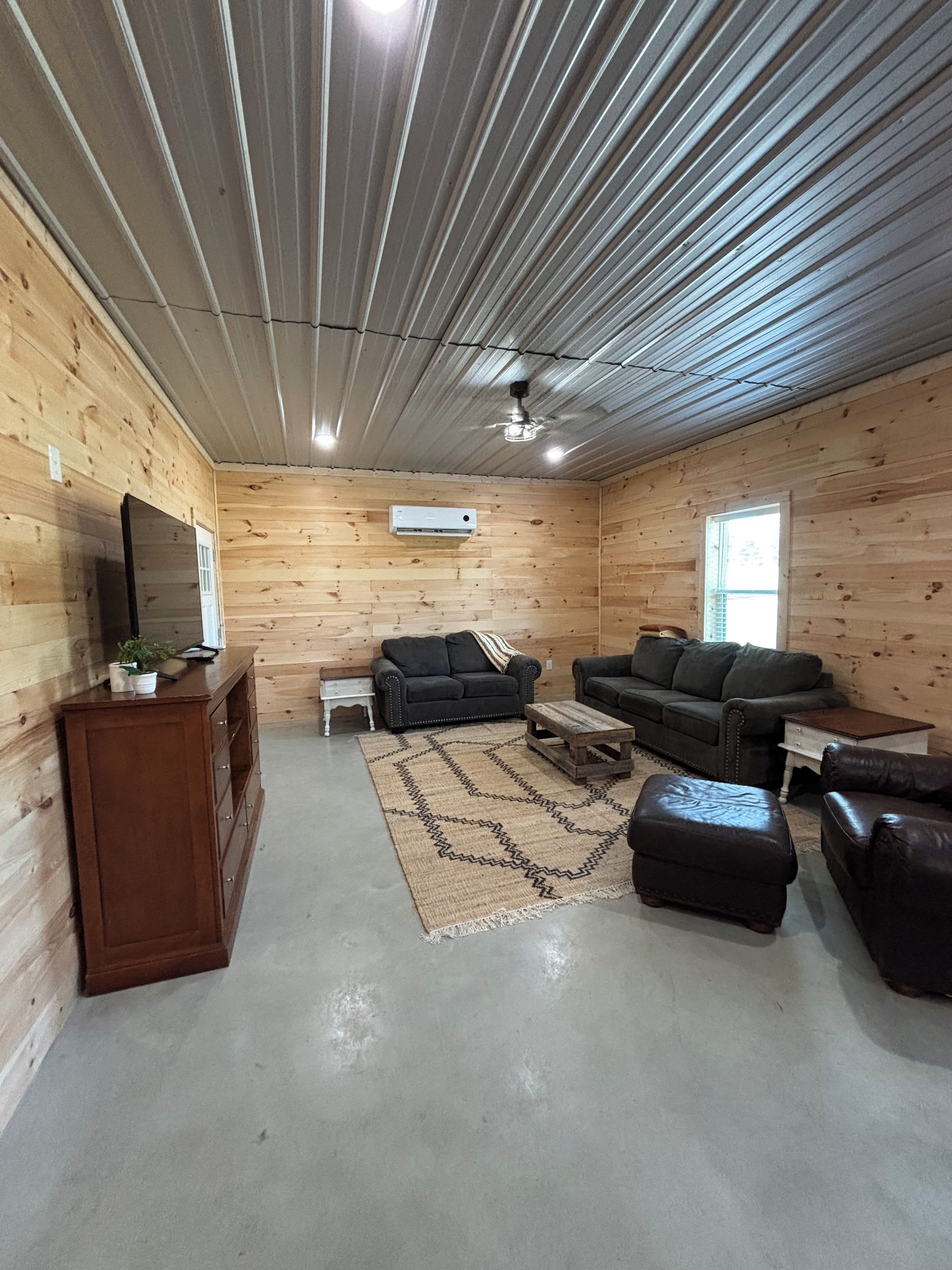 Interior de cabana de tronco com piso de madeira, ventilador de teto, televisão sobre um móvel de madeira e área de estar com sofá e apoio para os pés.