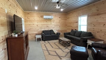 Interior de cabana de tronco com piso de madeira, ventilador de teto, televisão sobre um móvel de madeira e área de estar com sofá e apoio para os pés.