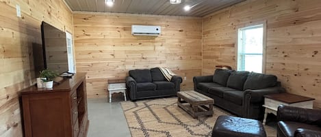 A log cabin interior with a wooden floor, a ceiling fan, a television on a wooden cabinet, and a seating area with a sofa and ottoman.
