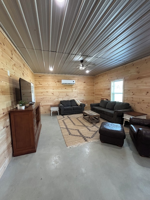 A log cabin interior with a wooden floor, a ceiling fan, a television on a wooden cabinet, and a seating area with a sofa and ottoman.