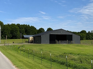 A large, dark-colored barn with an open door, surrounded by a fenced pasture and a clear sky.