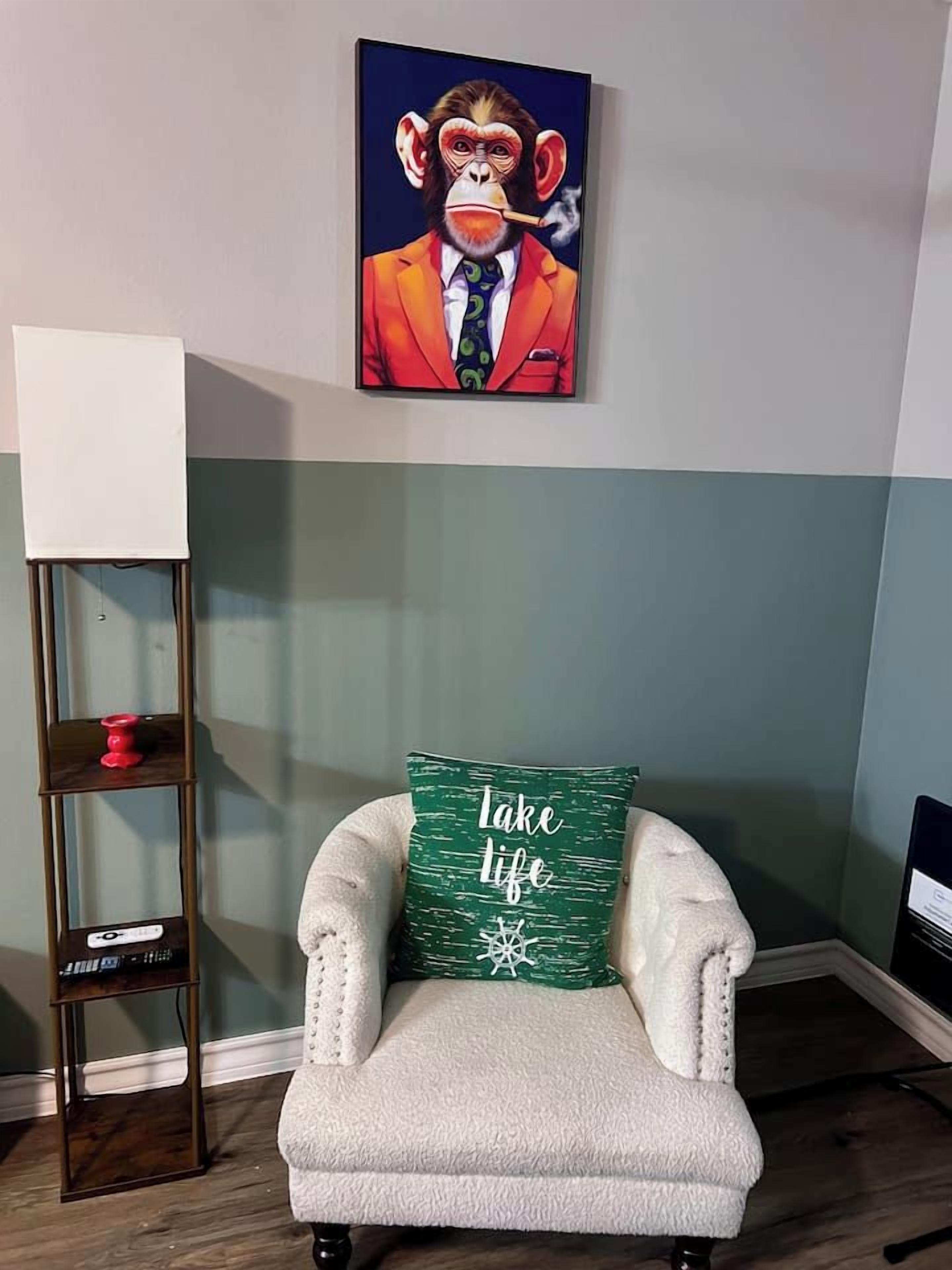 Ett rum med en vit fåtölj, en grön kudde med text på, en tavla på väggen och en hög golvlampa.