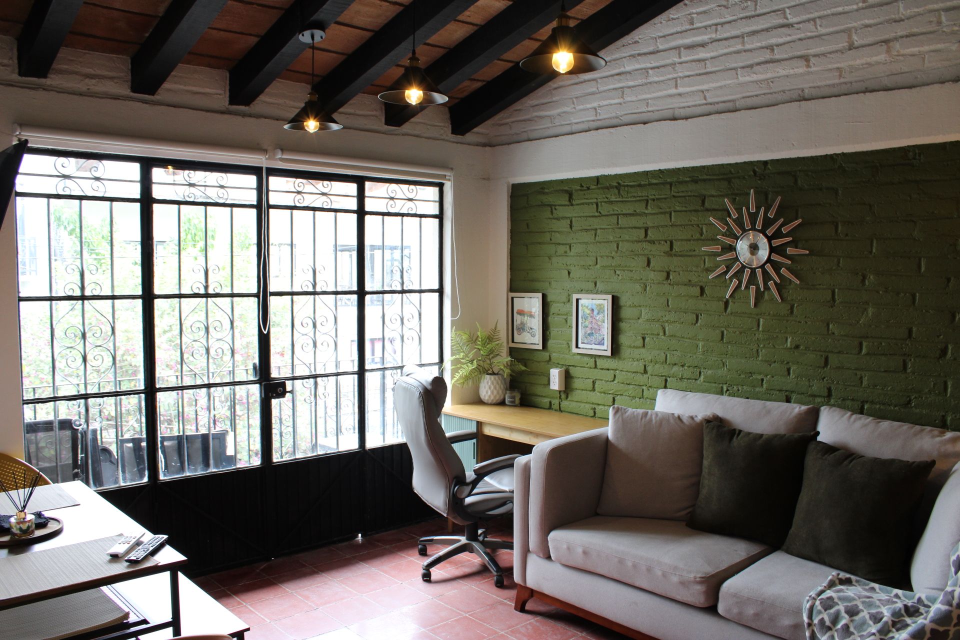 Una habitación con pared de ladrillo verde, un sofá blanco con cojines, un escritorio con una silla y una ventana con barrotes.