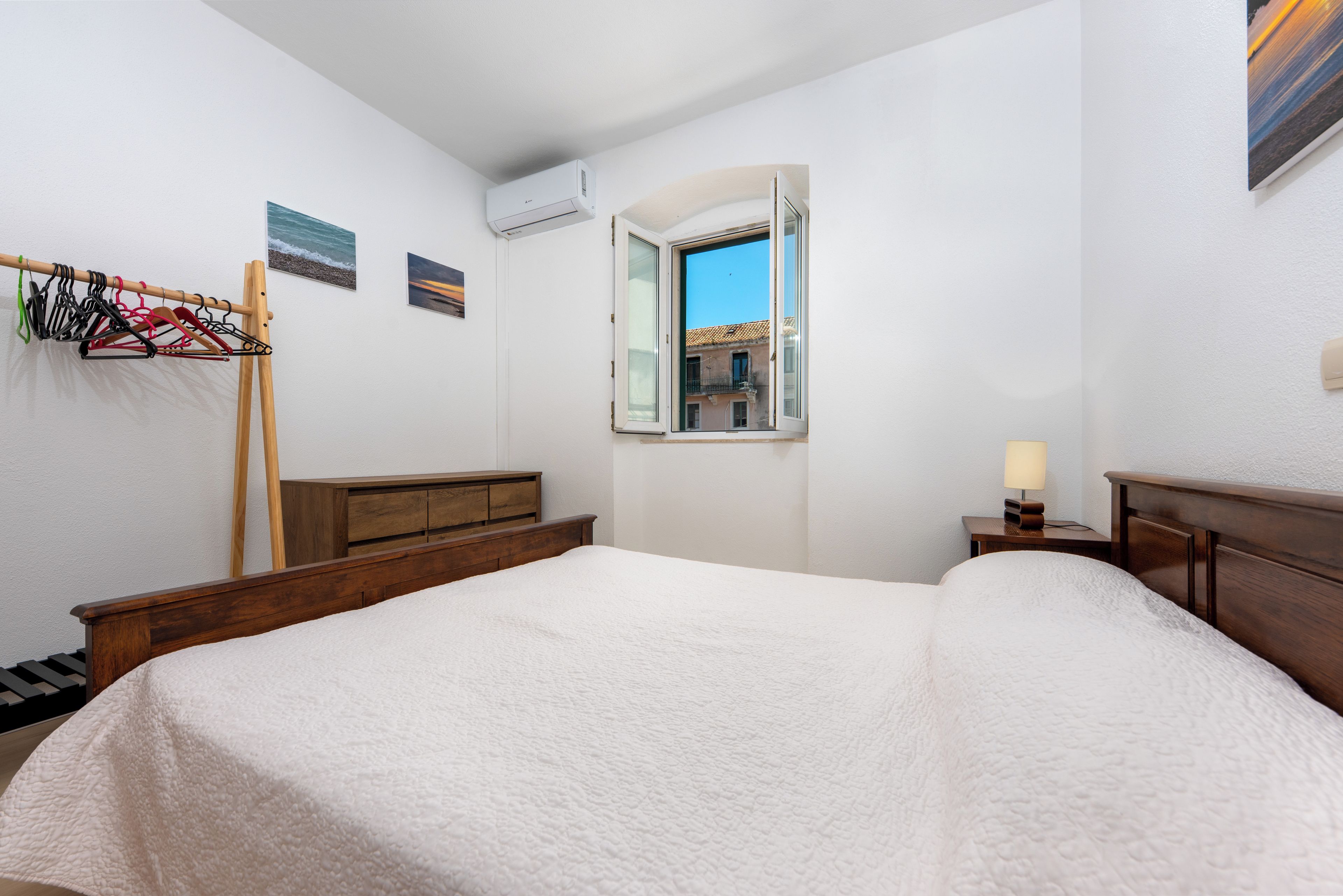 A hotel room with a bed, a wooden dresser, a window with a view of buildings, and a coat rack with hangers.