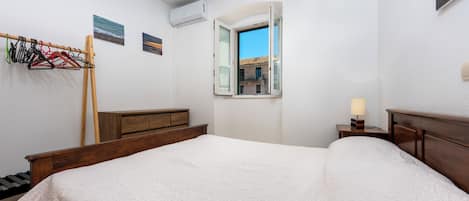 A hotel room with a bed, a wooden dresser, a window with a view of buildings, and a coat rack with hangers.