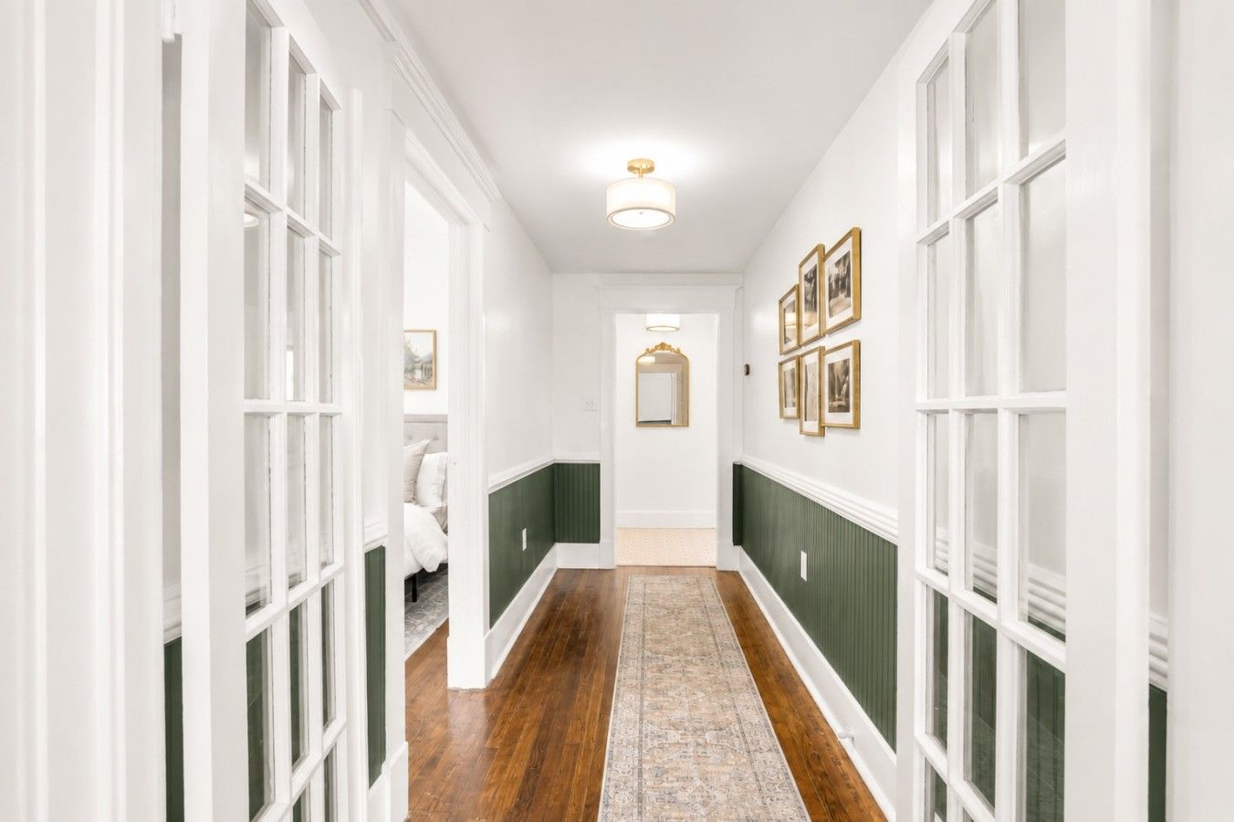 Een gang met witte wanden, groene lambrisering, houten vloeren en een vloerkleed. Er hangen ingelijste foto’s aan de muur en een spiegel.