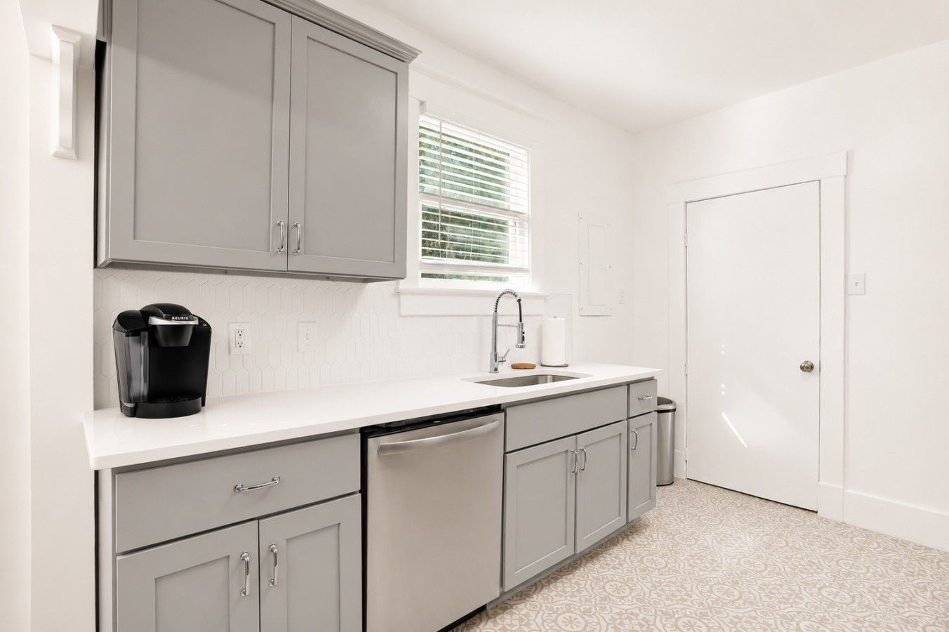 A compact kitchen with grey cabinets, a dishwasher, a coffee maker, and a window with blinds.
