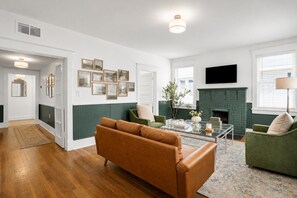 Un salon doté d’une cheminée, de fauteuils à revêtement vert, d’un canapé marron et d’un téléviseur à écran plat fixé au mur.
