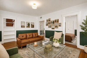 A living room with a brown leather sofa, a green armchair, a glass coffee table with decorative items, and a bookshelf filled with books. - Charming & Modern Savannah Retreat | Steps to Daffin Park (Savannah)