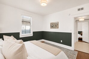 Un dormitorio con cama, una ventana con persianas, un cuadro en la pared y una puerta que da a otra habitación.