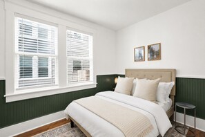 A bedroom with a bed, two bedside tables, a window with blinds, and two framed pictures on the wall. - Charming & Modern Savannah Retreat | Steps to Daffin Park (Savannah)