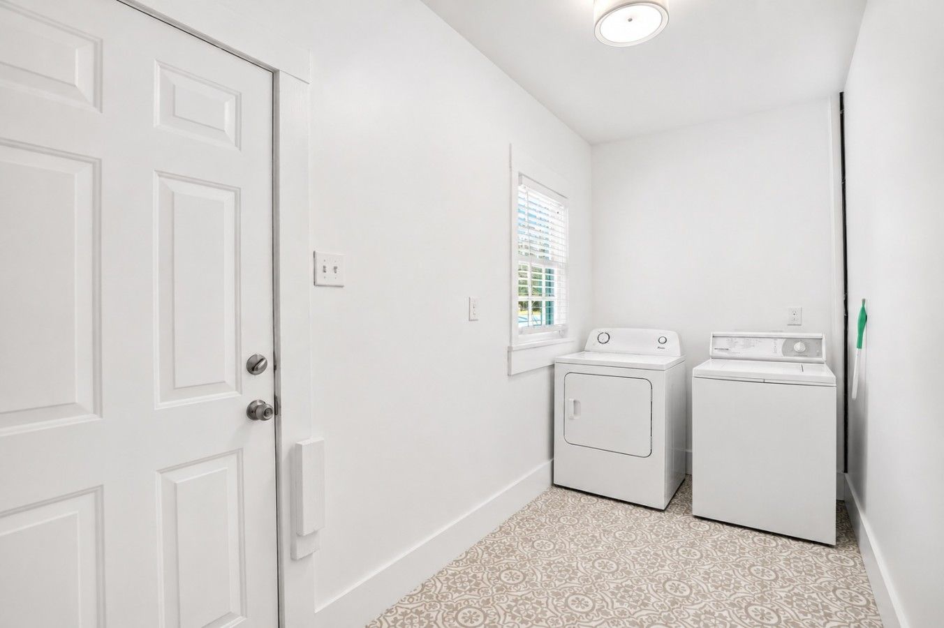 A laundry room with a door, a window with blinds, and two white appliances.