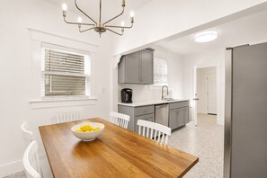 A modern kitchen with grey cabinets, a wooden dining table with chairs, and a bowl of fruit. - Charming & Modern Savannah Retreat | Steps to Daffin Park (Savannah)