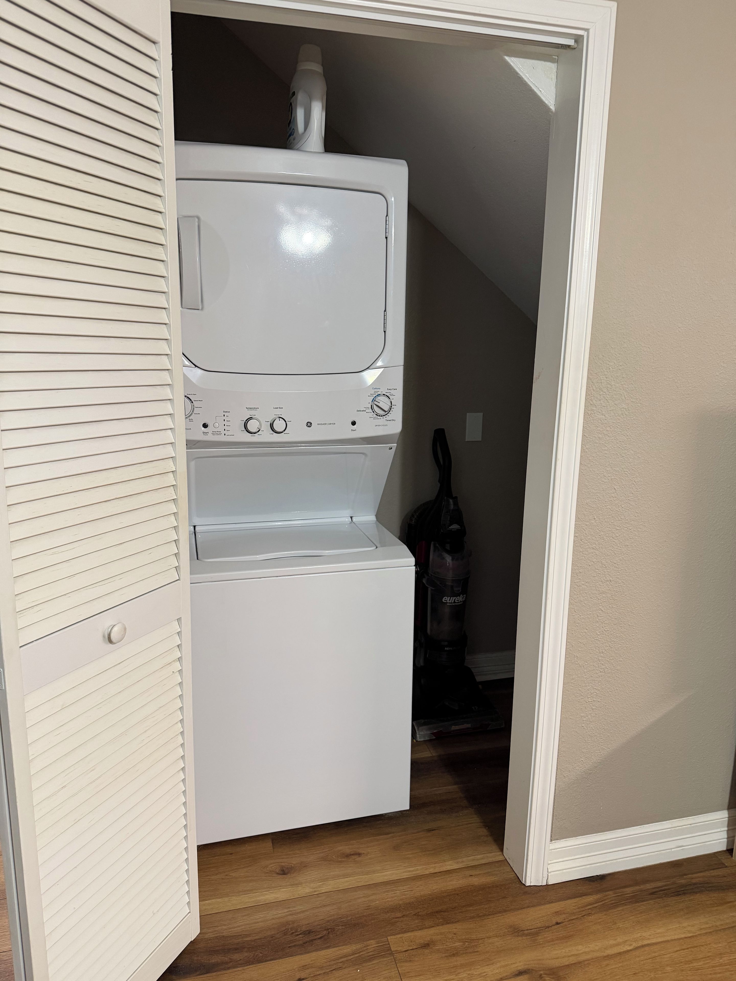 A laundry room with a stacked washer and dryer, a bottle of detergent, and a vacuum cleaner.