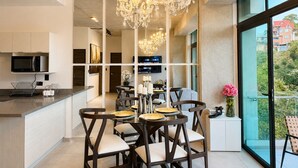 A modern kitchen with a central island, a dining area with dark wooden chairs and a chandelier, and a large window offering a view of greenery. - Park Tower | Santa Elena Sur Area & U.S. Embassy (Nuevo Cuscatlan)