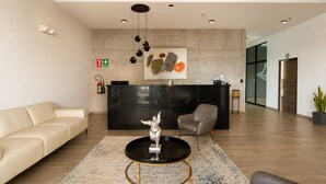 A modern reception area with a black reception desk, a beige sofa, a grey armchair, and a unique sculpture on a coffee table. - Park Tower | Santa Elena Sur Area & U.S. Embassy (Nuevo Cuscatlan)