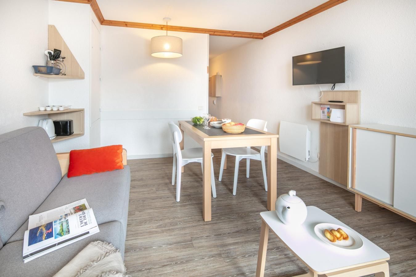 Ein modernes Wohnzimmer mit einem grauen Sofa, einem Esstisch mit Stühlen, einem Fernseher und einer kleinen Küchenzeile mit Wasserkocher und Toaster.