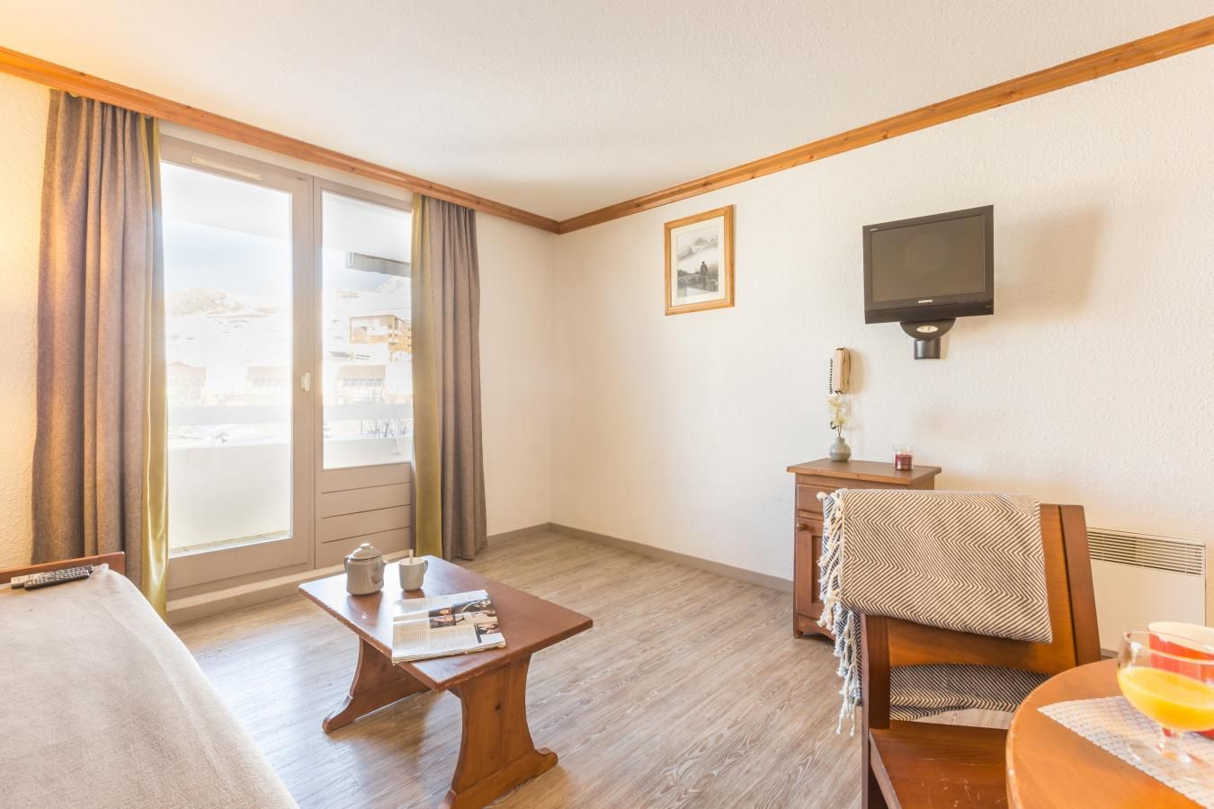 A hotel room with a bed, a small table with a newspaper, a chair, a television mounted on the wall, and a window with curtains.
