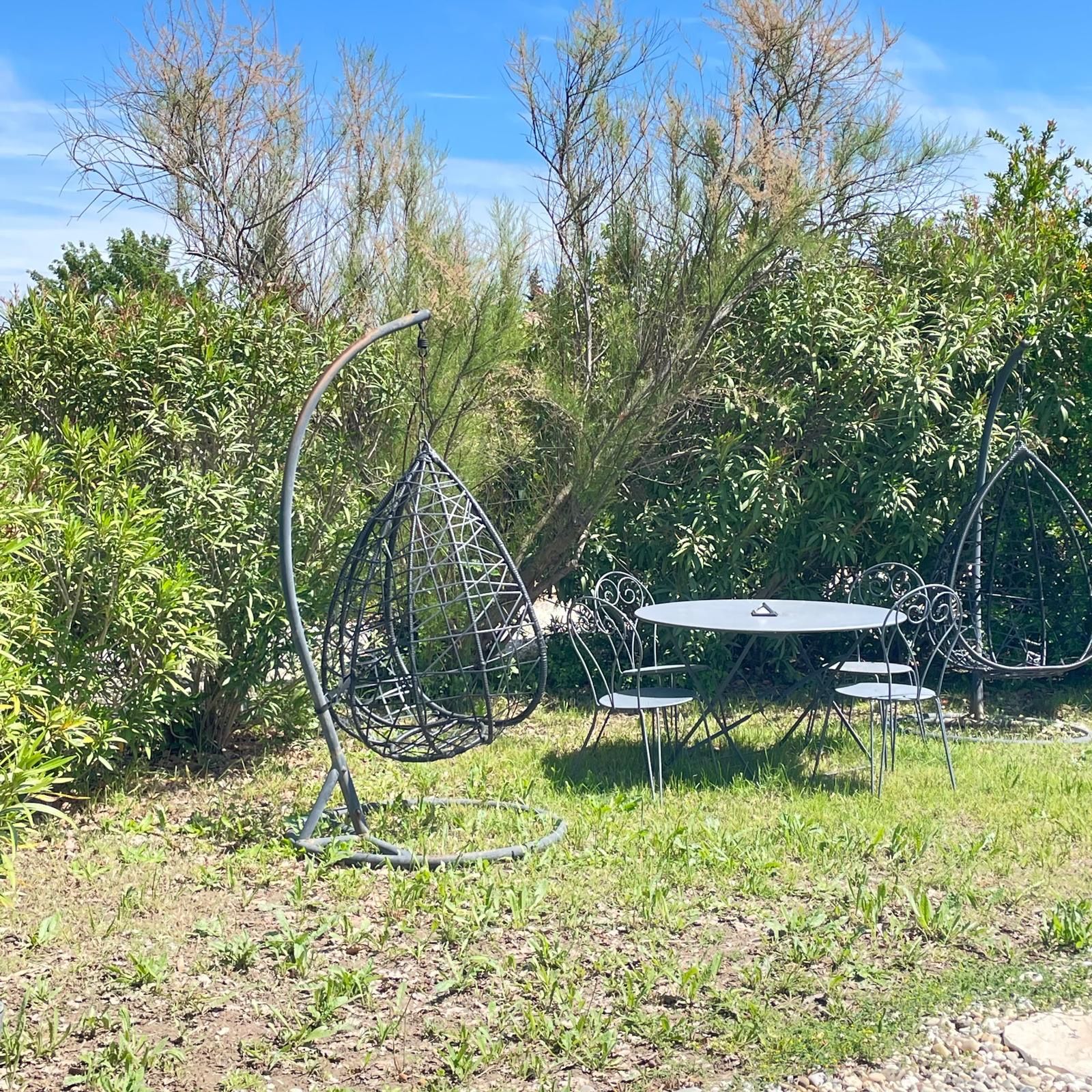 A metal outdoor seating area with a round table and chairs, surrounded by greenery and trees.