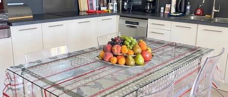 Une cuisine moderne avec des armoires blanches, une hotte de cuisinière en acier inoxydable et une table en verre agrémentée d’un centre de table composé de fruits.