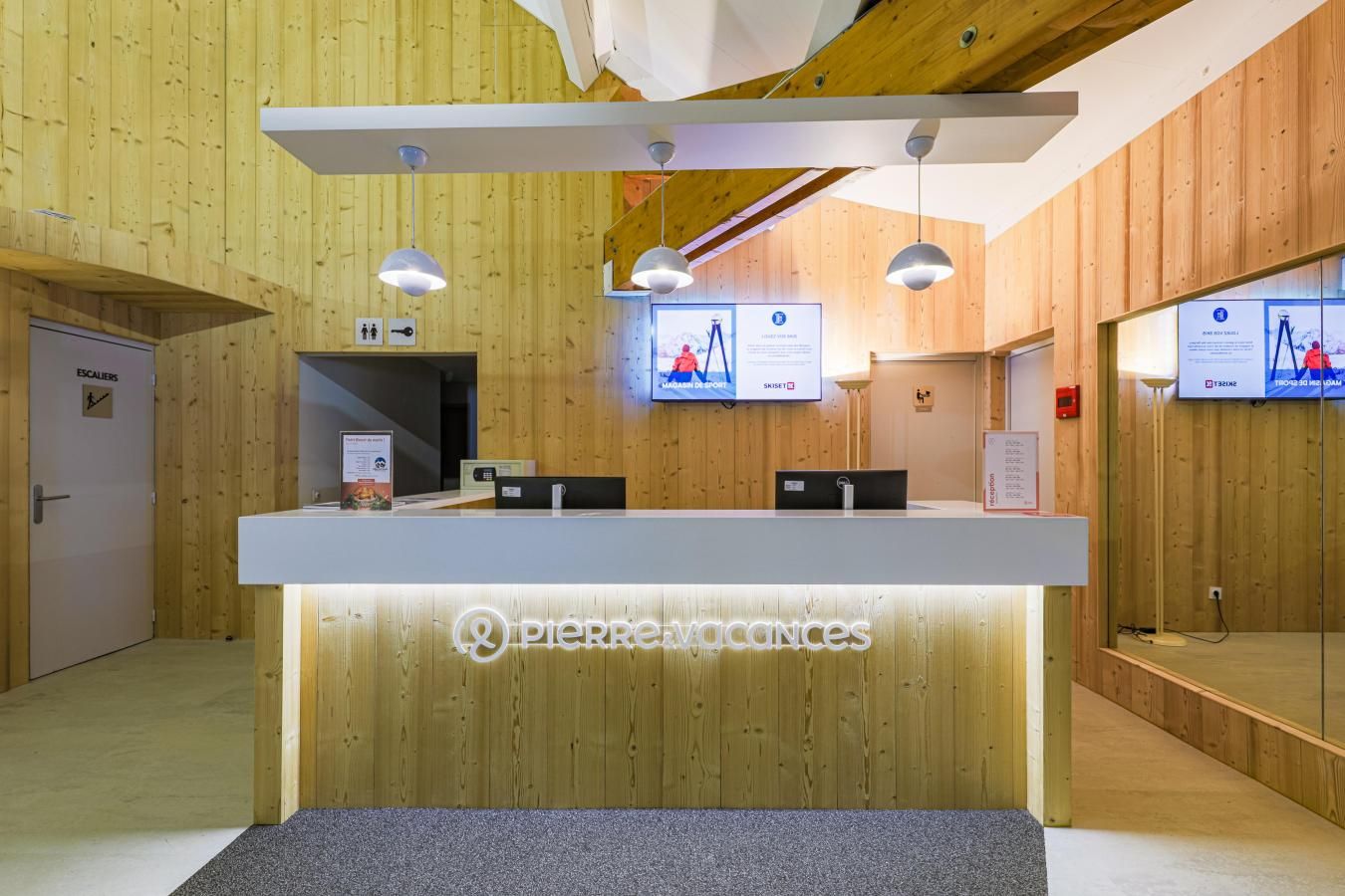 Una mesa de recepción con detalles de madera, una pantalla digital que muestra información y un cartel que indica la ubicación de los ascensores.